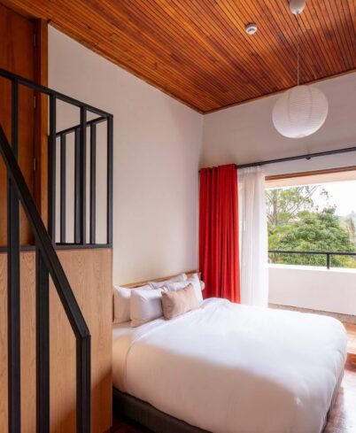 Cosy hotel room with double bed, balcony view, wooden decor, and red curtains welcoming natural light.